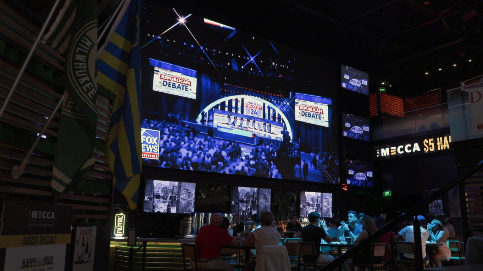 People sit at multiple tables underneath a large screen showing the FOX News Channel presentation of a presidential primary debate, in a room with multiple smaller television screens, a backlit sign reading The MECCA, and flags for the Milwaukee Bucks and Milwaukee Brewers.