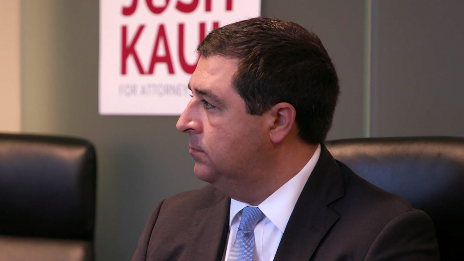 Josh Kaul is seen in profile while seated in a high-backed chair in a room with an out-of-focus campaign sign for his 2022 candidacy in the background.