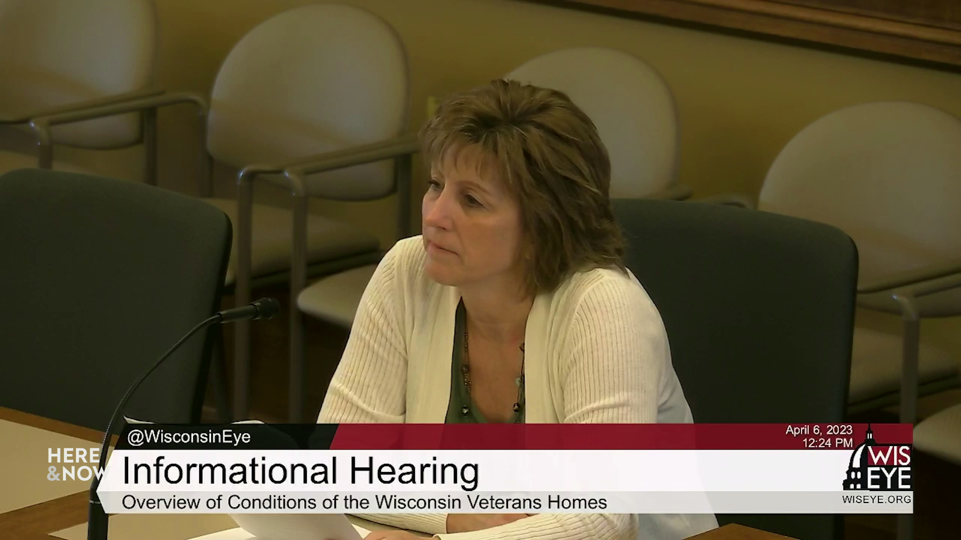 A video still image shows Laurie Miller sitting at a table while speaking into a microphone in a room with multiple empty chairs in the background, with a video graphic at bottom including the text "Informational Hearing" and "Overview of Conditions of the Wisconsin Veterans Homes."
