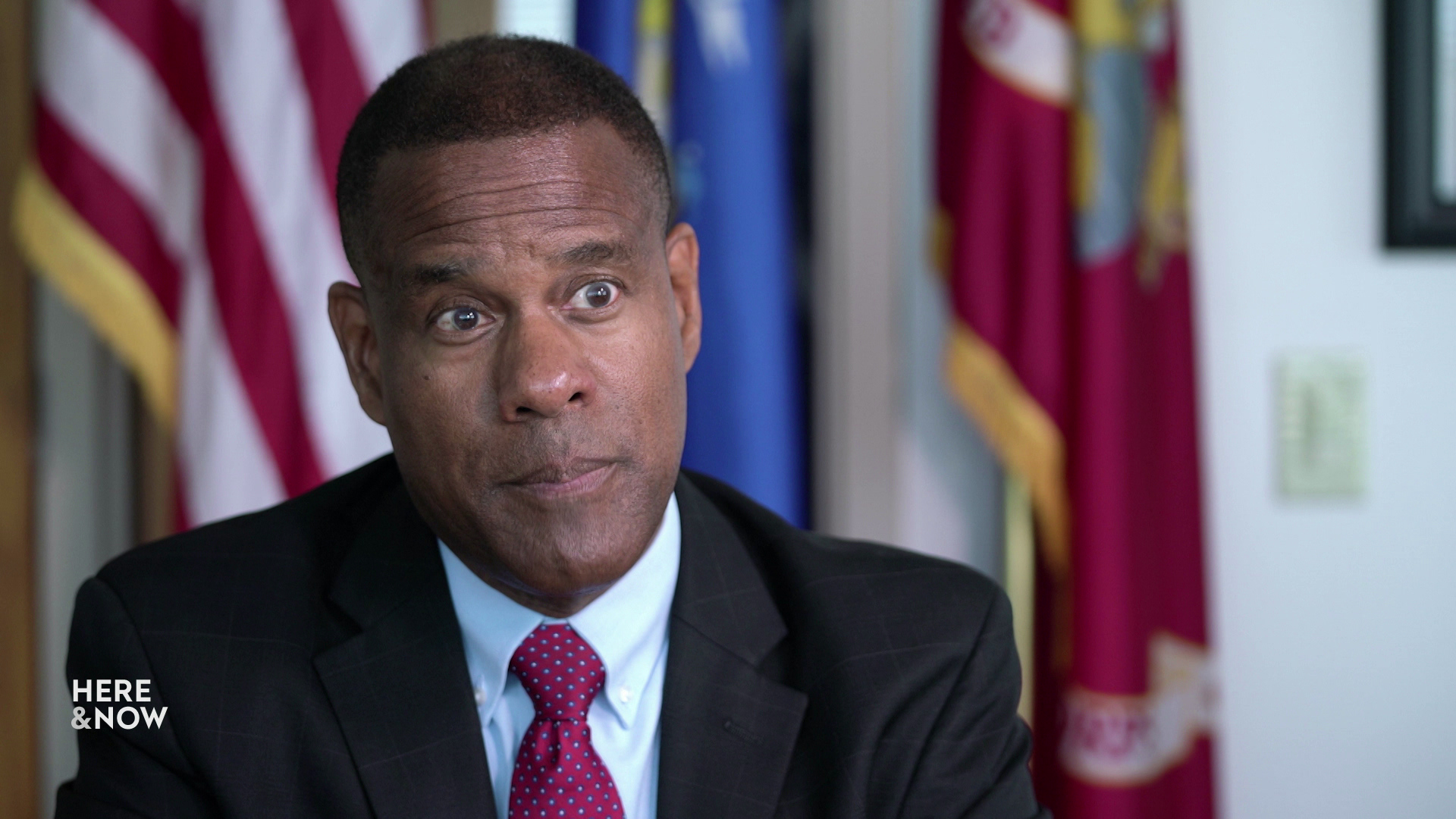 A still image from an interview shows James Bond sitting in a room with the U.S. and U.S. Armed Forces branch flags in the background.