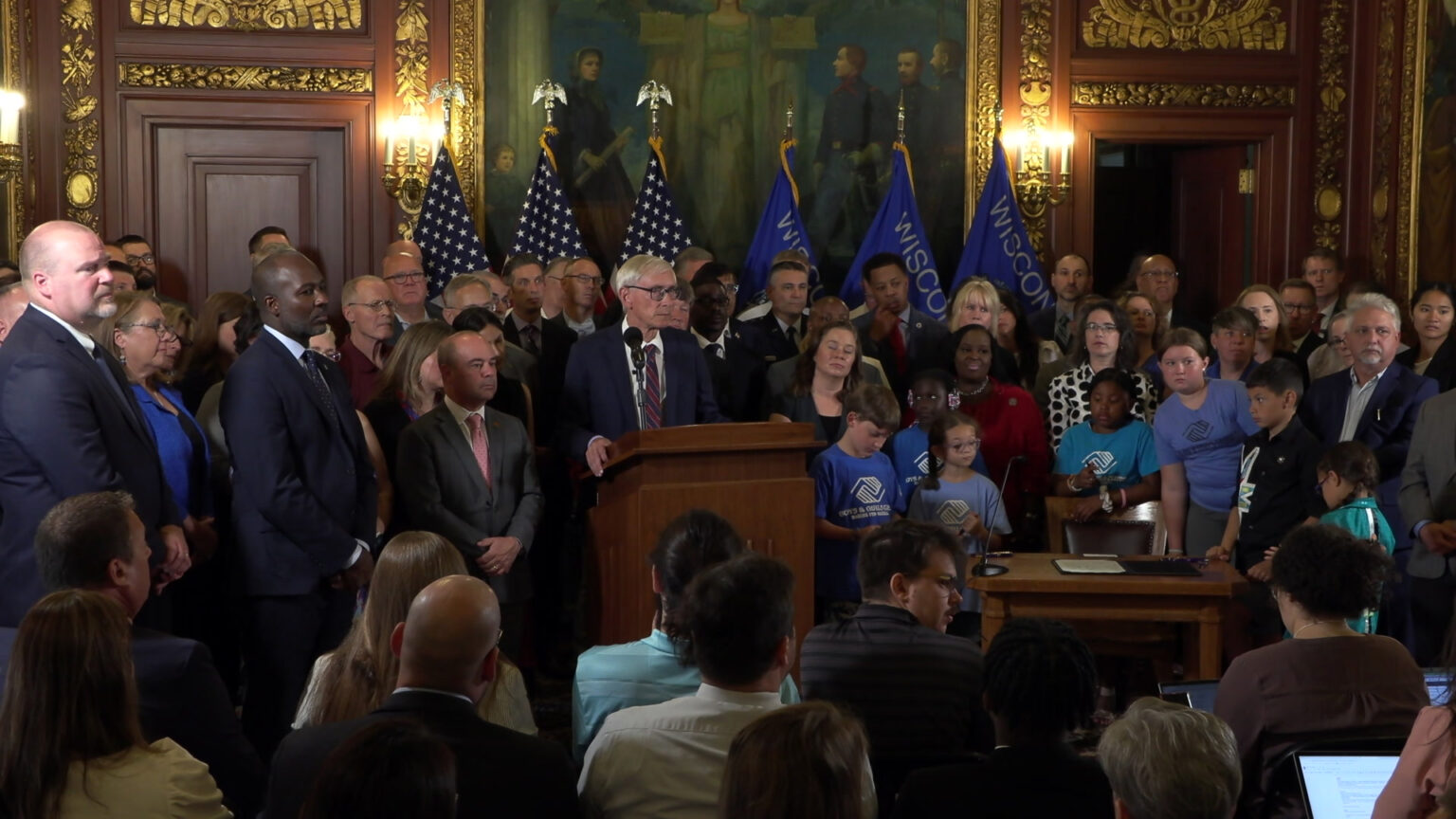 Tony Evers stands and speaks into a microphone mounted on a wood podium, with people standing behind him and to his side, and more people seated facing him, in a room with U.S. and Wisconsin flags, a painting, electric wall sconce lighting, and gold-colored filigree décor framing two doors.