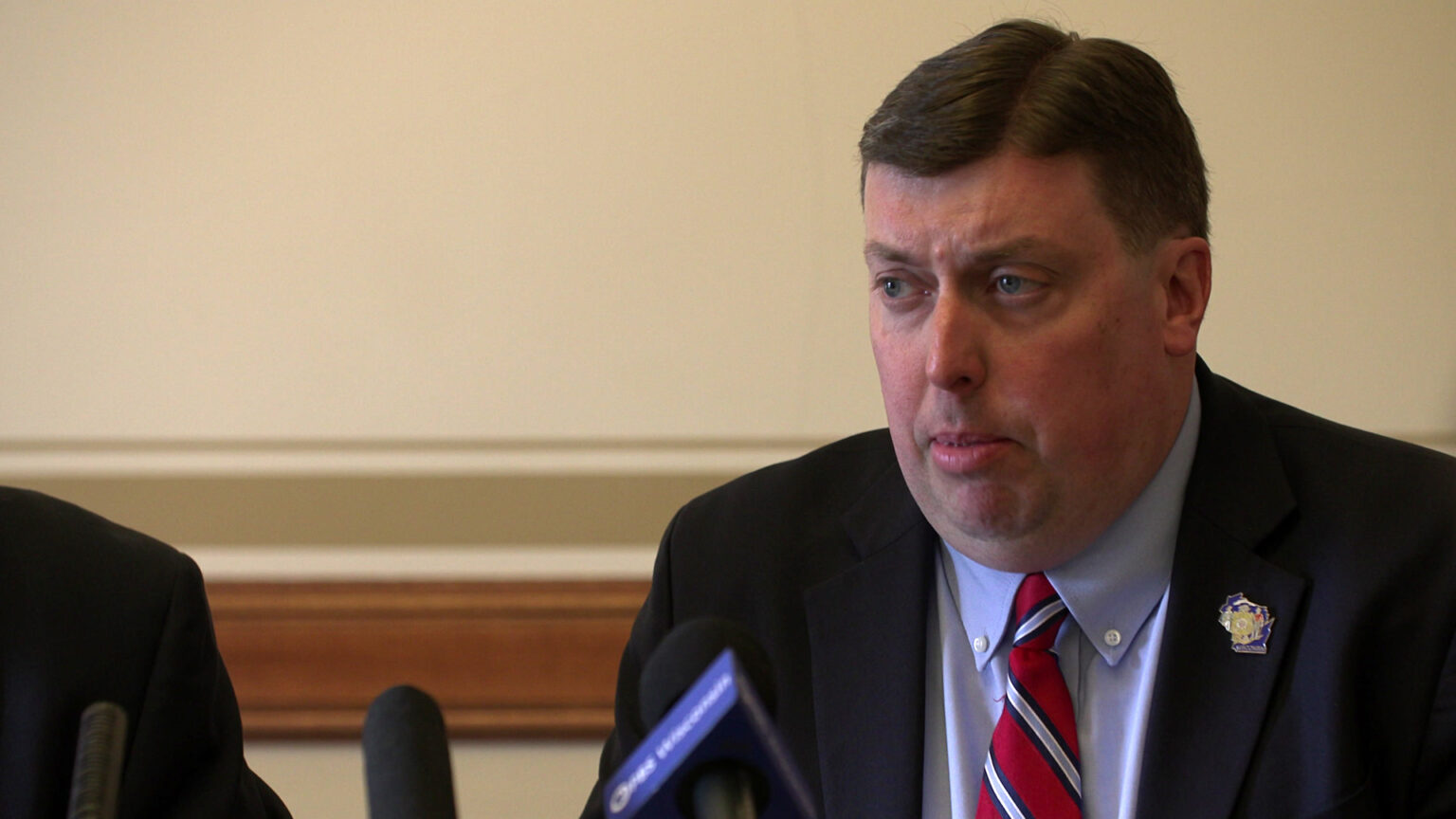 Mark Born speaks into multiple microphones while seated in a room with wood wainscoting trim in the background.