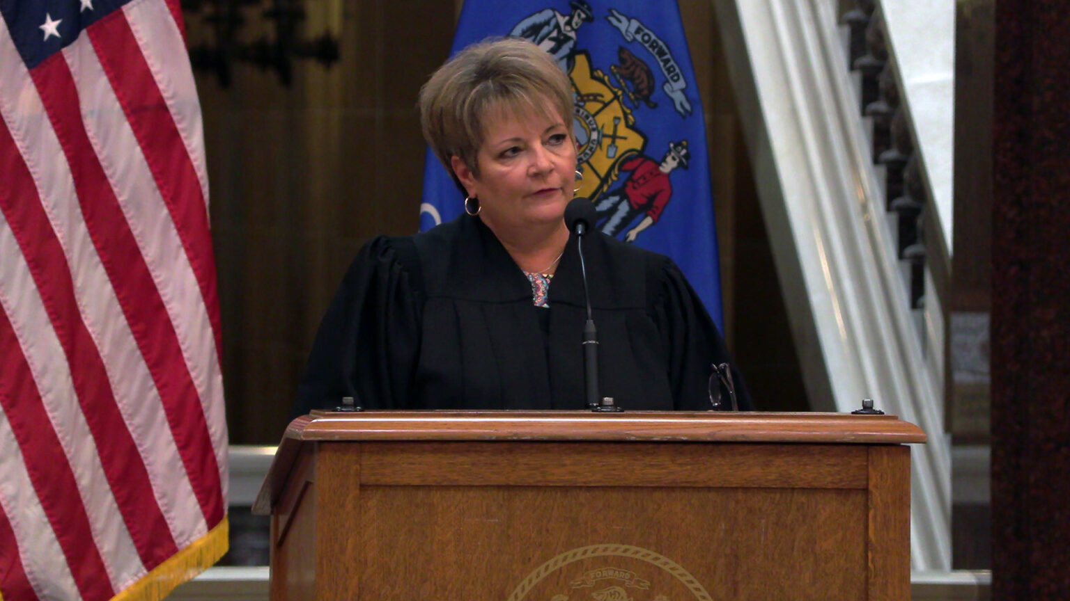 Janet Protasiewicz stands and speaks into a microphone mounted on a wood podium, with the U.S. and Wisconsin flags, as well as a marble staircase balustrade in the background. 
