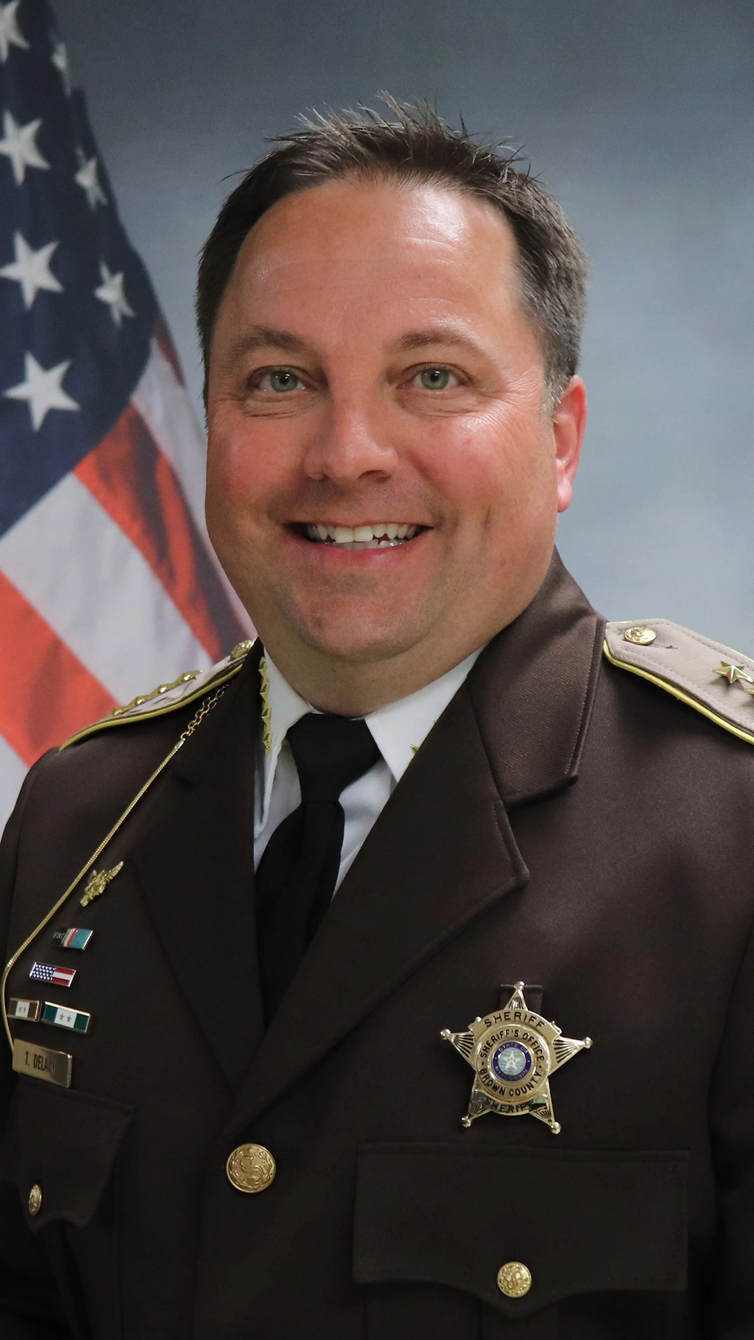 Todd Delain poses for a portrait while wearing a sheriff's uniform and with a U.S. flag on a graphic in the background.