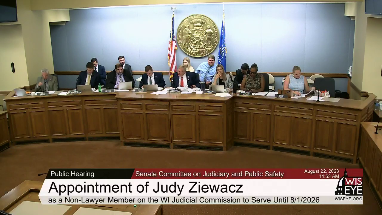 A video still image shows eight people sitting at a legislative dais in front of microphones and nameplates, with other people seated in chairs behind, in a room with the U.S. and Wisconsin flags on either side of the Great Seal of the State of Wisconsin mounted on the rear wall, with a video graphic at bottom including the text Appointment of Judy Ziewacz as a Non-Lawyer Member on the WI Judicial Commission to Serve Until 8/1/2026.