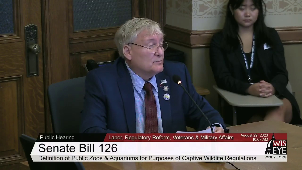 A video still image shows Van Wanggaard sitting at a table while speaking into a microphone in a room with another person seated in the background, with a video graphic at bottom including the text Senate Bill 126 and Definition of Public Zoos & Aquariums for Purposes of Captive Wildlife Regulations.