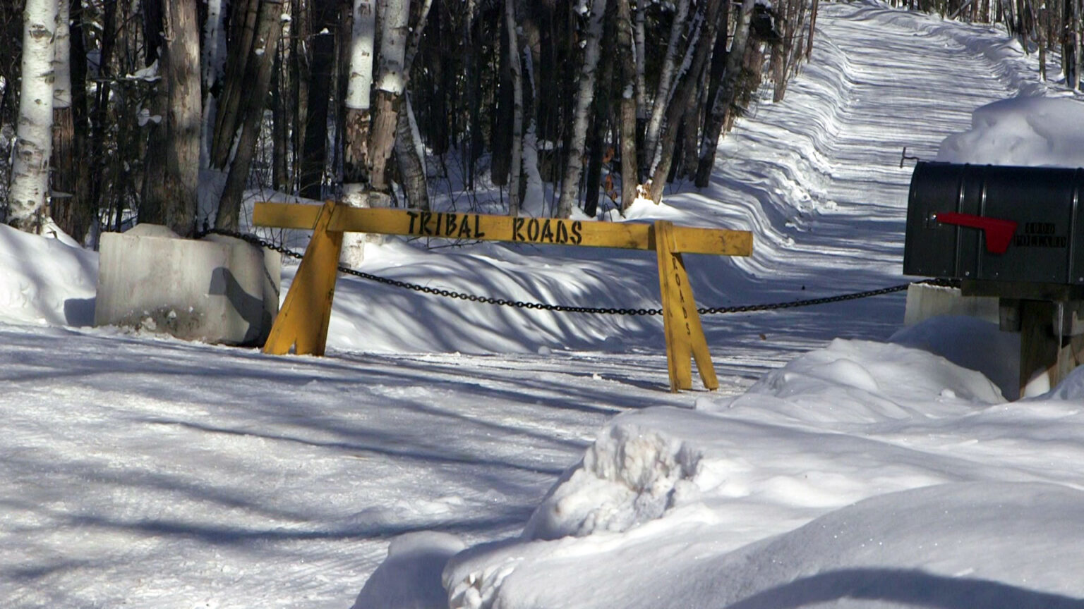 A wood road barrier with tribal roads painted on it and two concrete blocks connected with a metal chain sit in front of a road covered in snow, with a mailbox in the foreground and trees in the background.