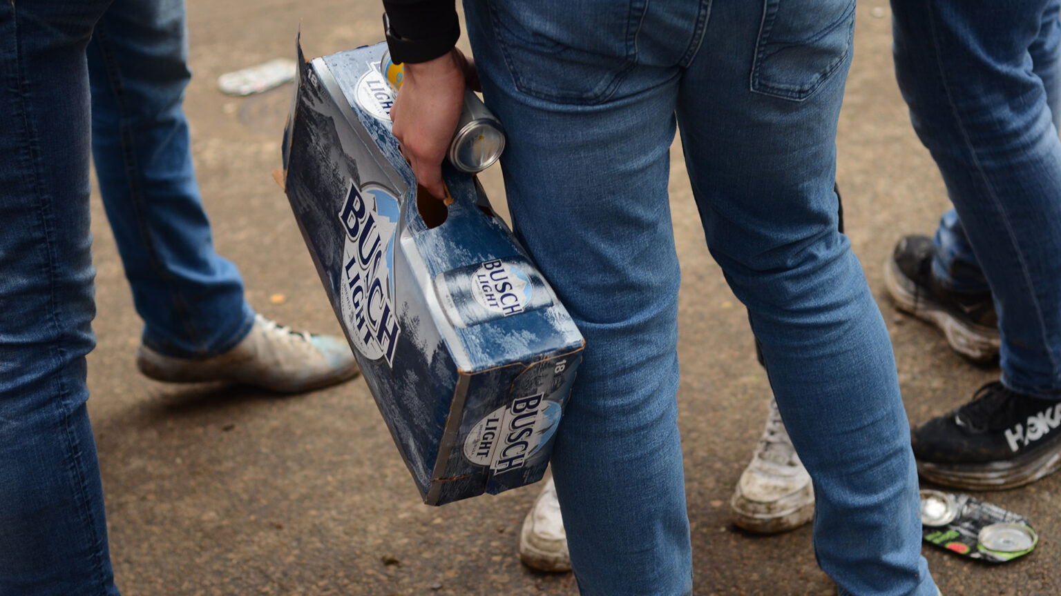 A person standing on pavement among other people holds a can of beer and a cardboard carton of beer cans in their left hand, with multiple crushed cans on the ground.