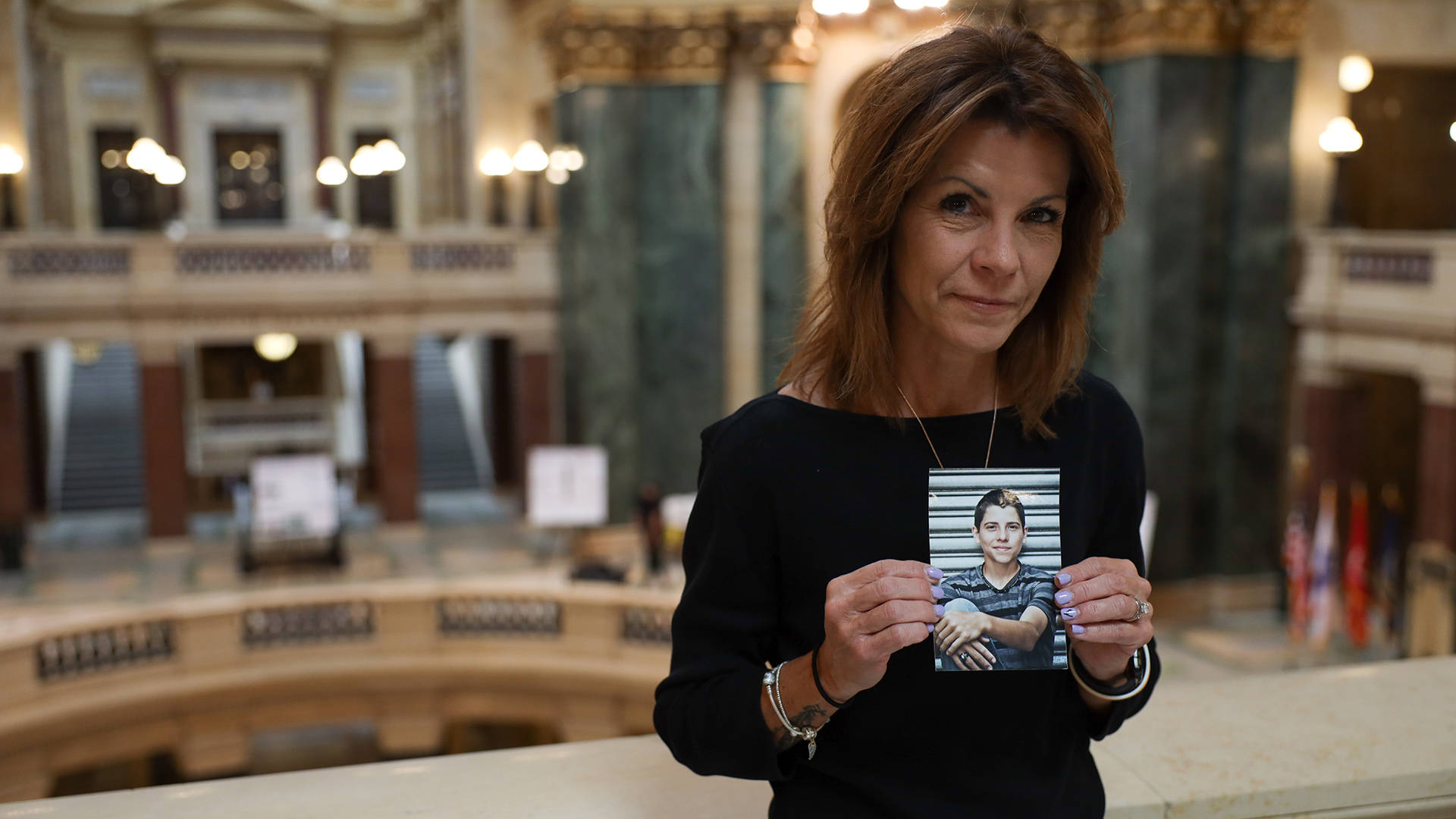 Sheila Lockwood poses for a portrait while holding a printed portrait of her son while standing at a marble railing overlooking a multiple levels rotunda, with walkways, stairs marble pillars and lights in the background.