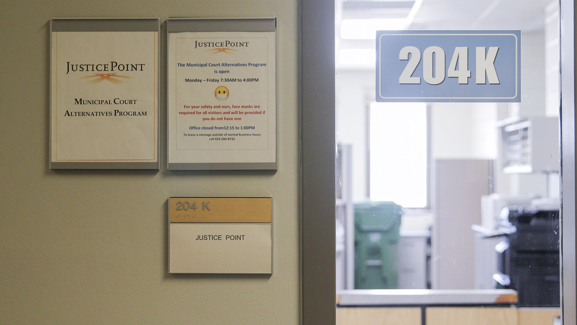 An office with a photocopy machine, shelves and other furniture is visible through a glass door window with a label reading "204K"and other signs reading "JusticePoint Municipal Court Alternatives Program" and showing its hours.
