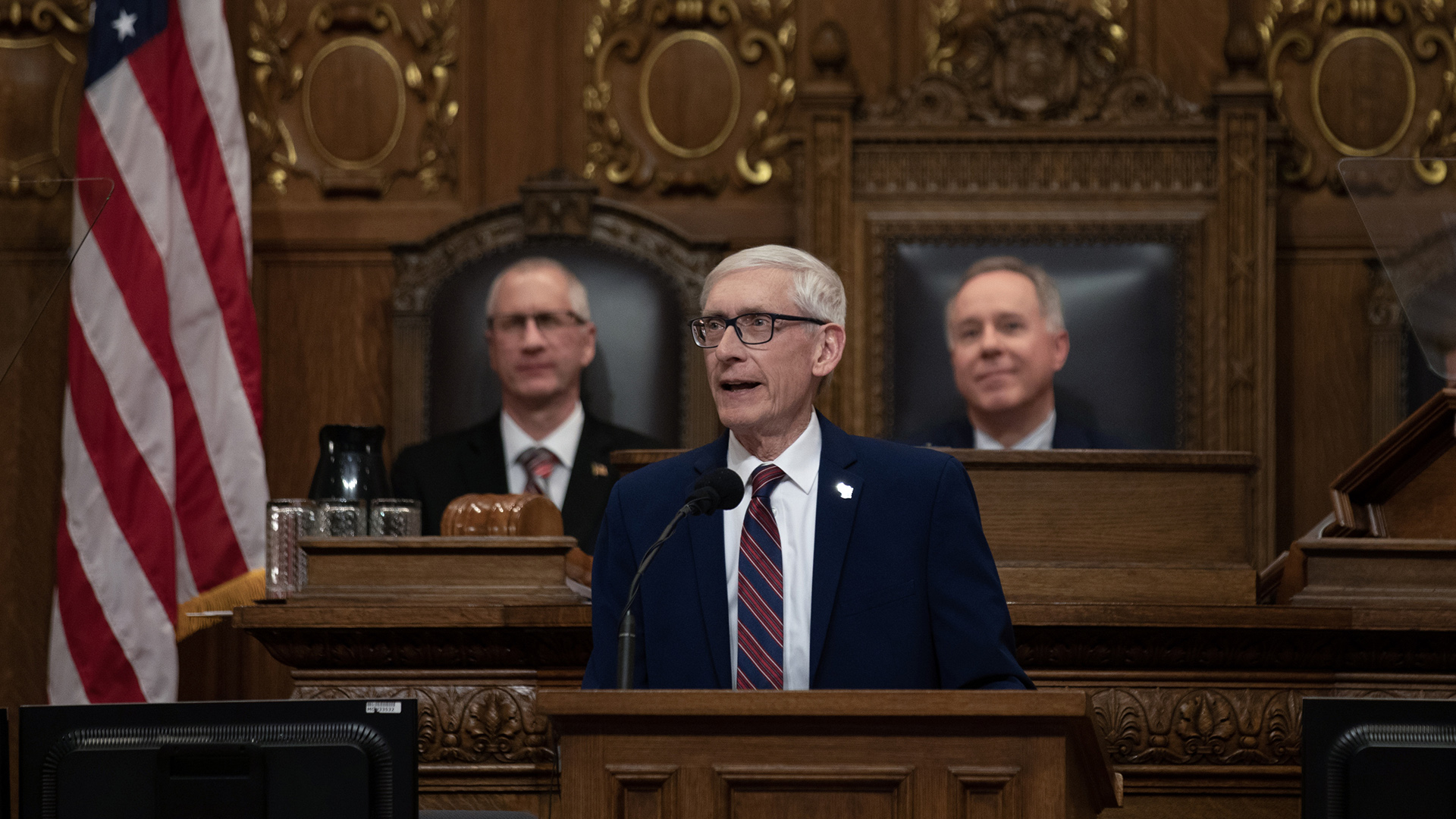 Tony Evers speaks into a microphone while standing behind a wood podium in front of a legislative dais with two people seated in high-backed wood and leather chairs, with a U.S. flag to his right, the backs of two monitors in the foreground, and carved wood wall paneling in the background.