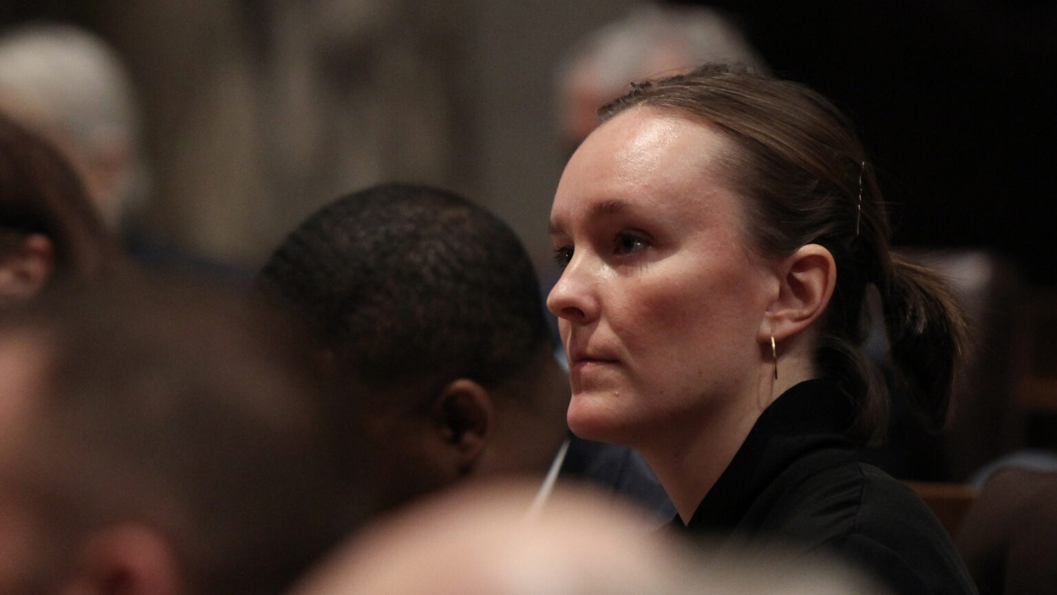 Greta Neubauer listens to a speaker while seated in a room among other people in the foreground and background.