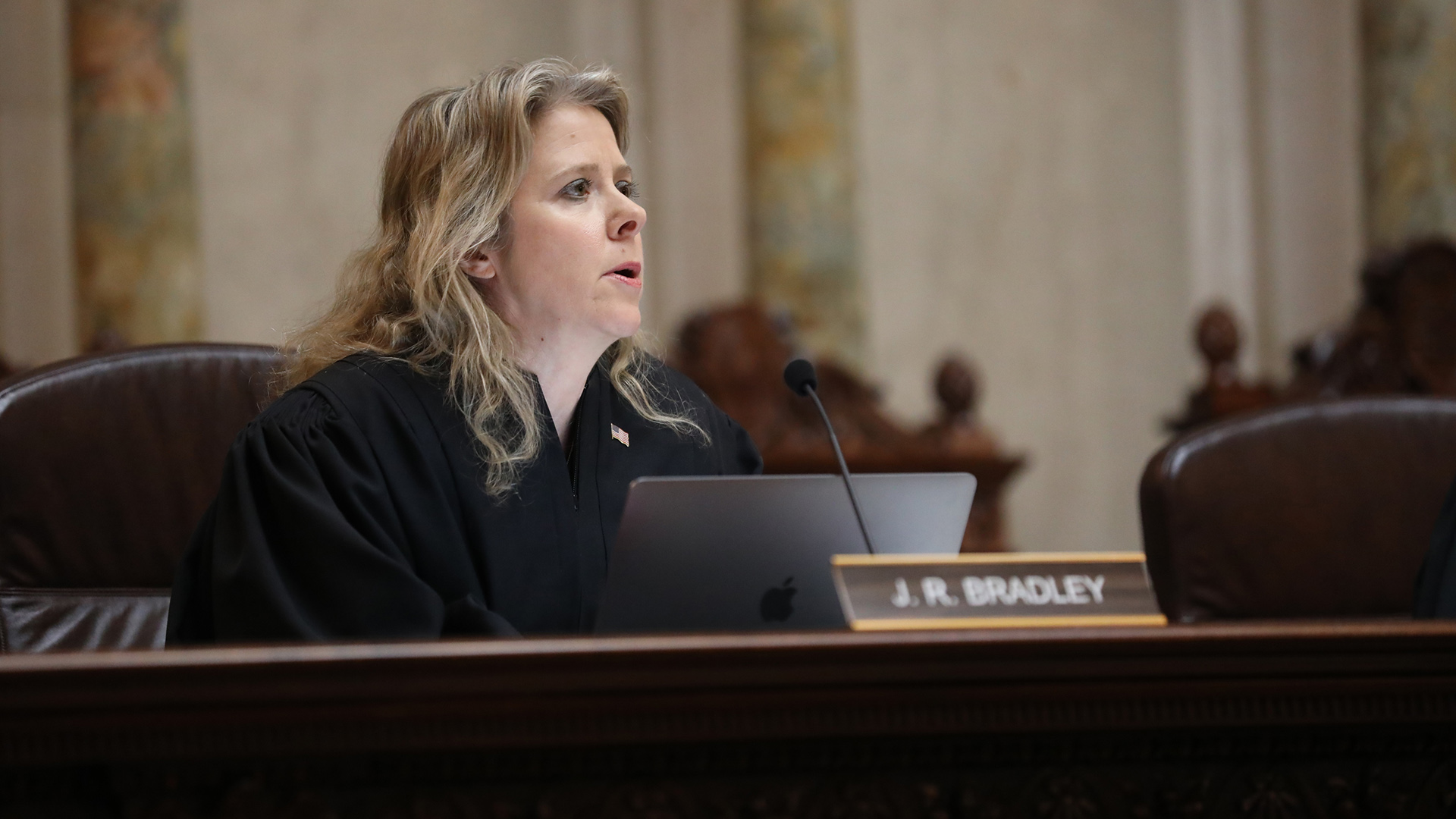 Rebecca Bradley sits in a high-backed leather chair behind a judicial bench and speaks into a mounted microphone while facing an open laptop computer, in a room with marble masonry.