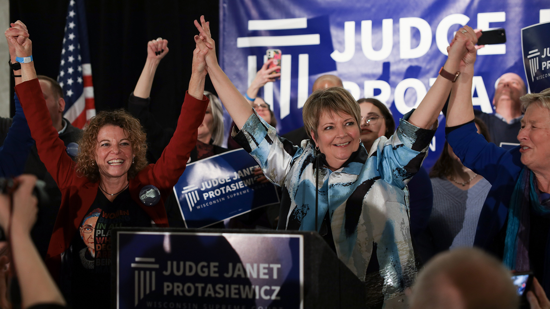 Janet Protasiewicz stands between Rebecca Dallet and Ann Walsh Bradley as they clasp hands and raise their arms in celebration, in a room with cheering people, a U.S. flag and multiple campaign signs that read "Justice Janet Protasiewicz,"