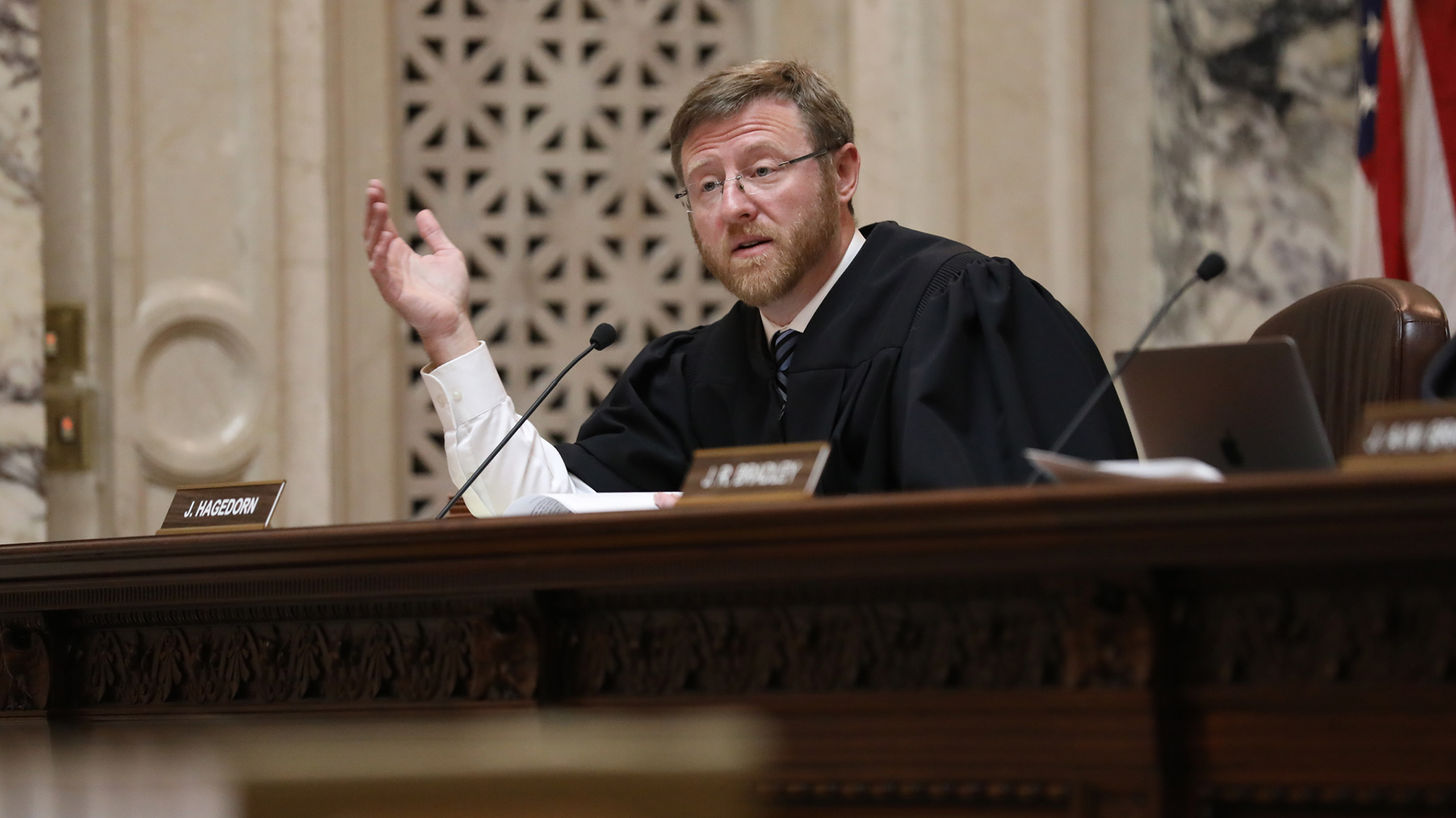 Brian Hagedorn sits at a judicial bench and gestures with his right hand while speaking into a table-mounted microphone, with papers and an open laptop computer to his side, in a room with a U.S. flag and marble masonry.