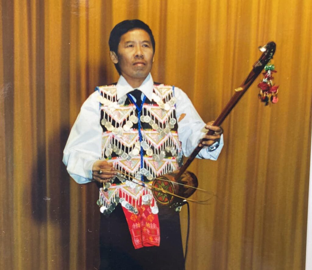 A man stands in front of a yellow curtain, playing the xim xaus, a traditional Hmong violin.