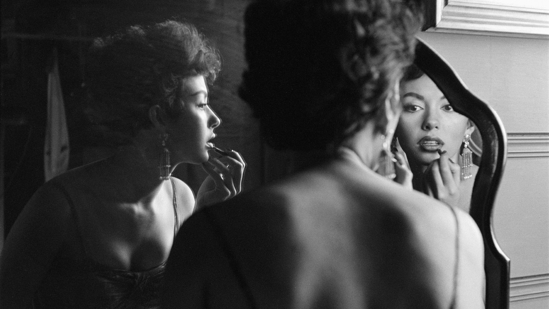 Black and white image of a woman looking into a mirror while applying lipstick.
