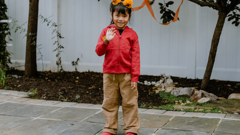 A child stands outside, smiling and waving at the camera, dressed up as Daniel Tiger.