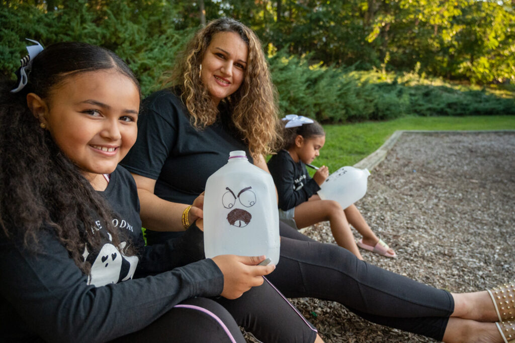 A parent and two children sit outside. The two children have homemade ghost decorations made from empty milk jugs.