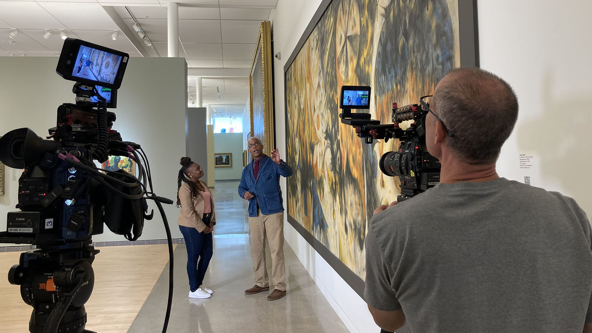 A woman interviews a man who discusses an art mural on a wall. There are video cameras recording the interaction.