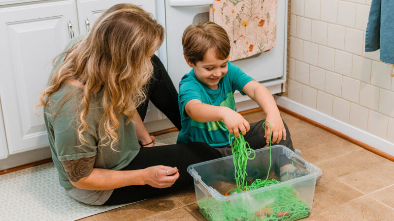 A parent and child play with the contents in a sensory bin, including green spaghetti noodles.