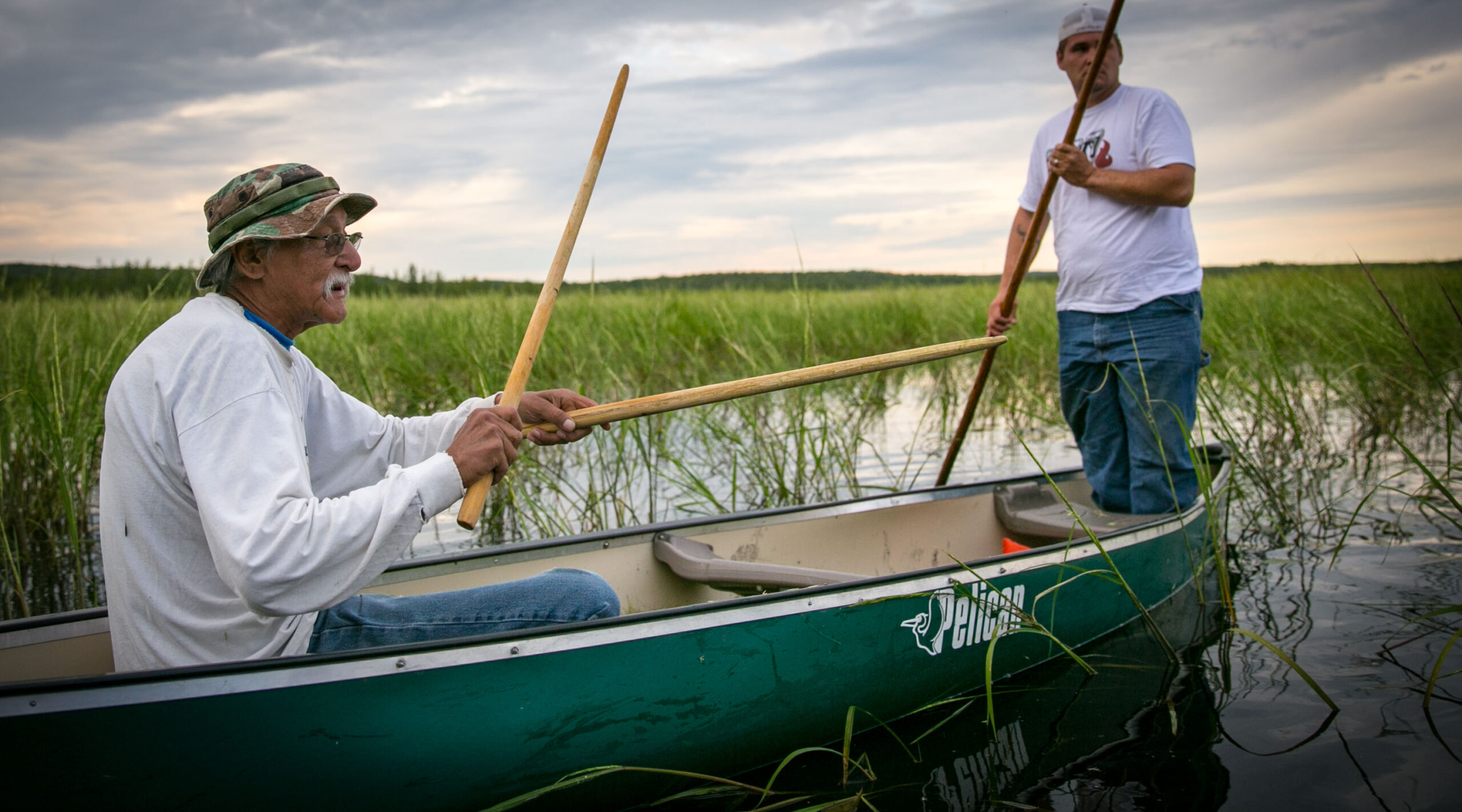 Two men ricing in a green canoe through a marshy area with tall grasses under a cloudy sky.