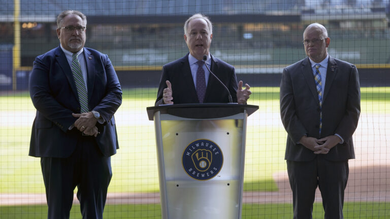 Robin Vos stands and gestures with both hands while speaking into a microphone affixed to a metal podium with the Milwaukee Brewers logo on its front, with Robert Brooks and Dan Feyen standing on either side, in front of protective netting and with a baseball infield and left-field wall and stands in the background.
