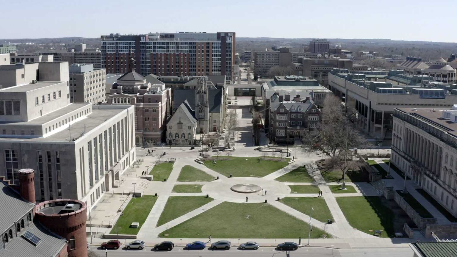 An aerial photo shows a plaza with sections of grass and concrete walkways surrounded by buildings of different sizes on three sides, with another walkway extending from its center toward the horizon between more buildings and trees.