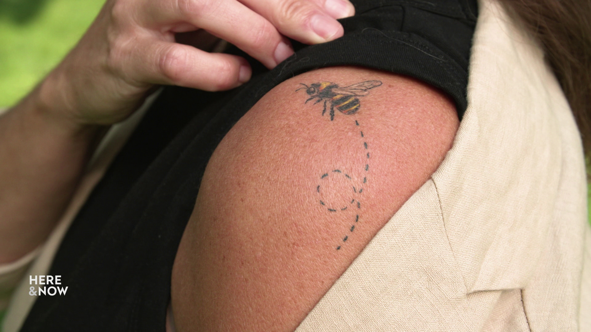 A hand lifts the edge of a shirt to show a yellow-and-black tattoo of a rusty patched bumble bee with a looping dotted line trailing from its abdomen on a shoulder.