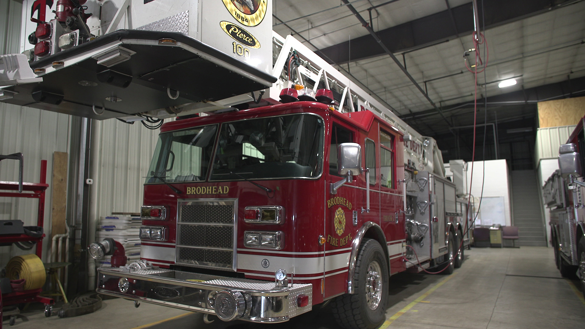 A fire truck with a roof-mounted latter sits parked inside an industrial garage with metal walls and an angled roof.