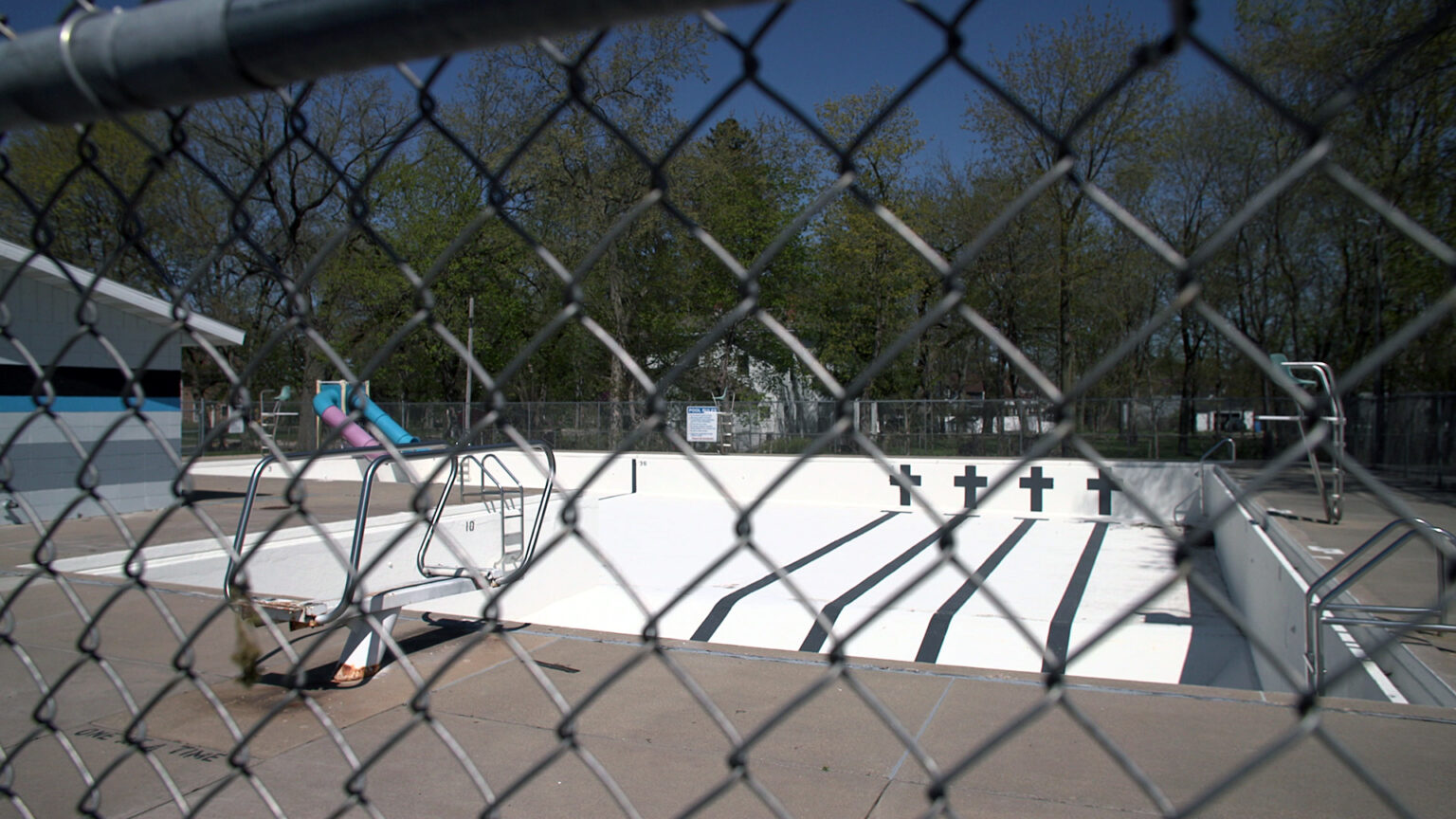 An empty pool with multiple swim lanes, a deeper diving area, and a shallower play area with two water slides sits empty, as seen through a chain-link fence, with buildings and trees in the background.
