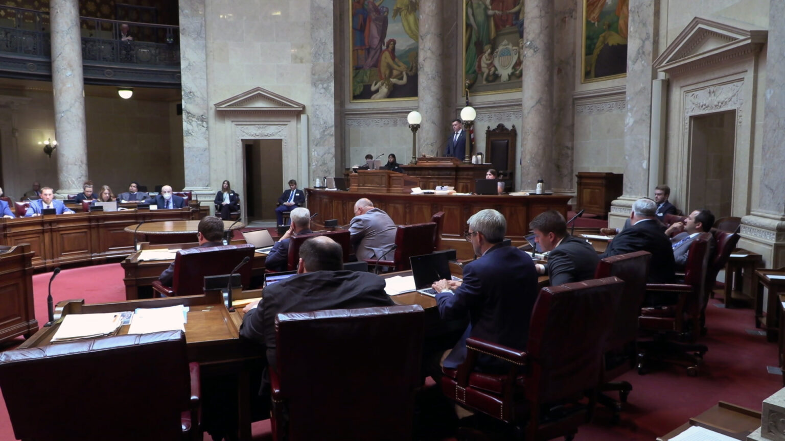 People sit in multiple, curved rows of desks equipped with microphones and look at laptop computers and speak to each other, in a room with marble masonry and pillars, large paintings, and a second-level seating gallery.