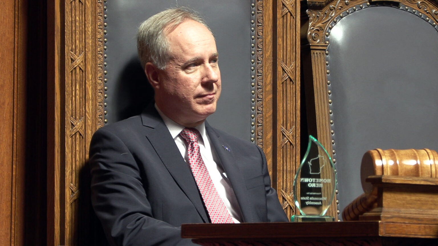 Robin Vos sits in a high-backed leather and wood chair and faces a desk with a glass award and large wood gavel, with another empty chair to his side.