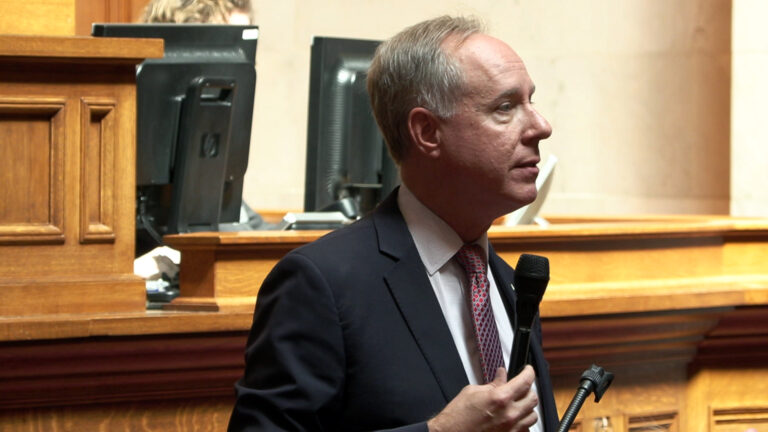 Robin Vos speaks into a microphone while standing in front of a wood legislative dais, with the back of two computer monitors on its surface in the background.