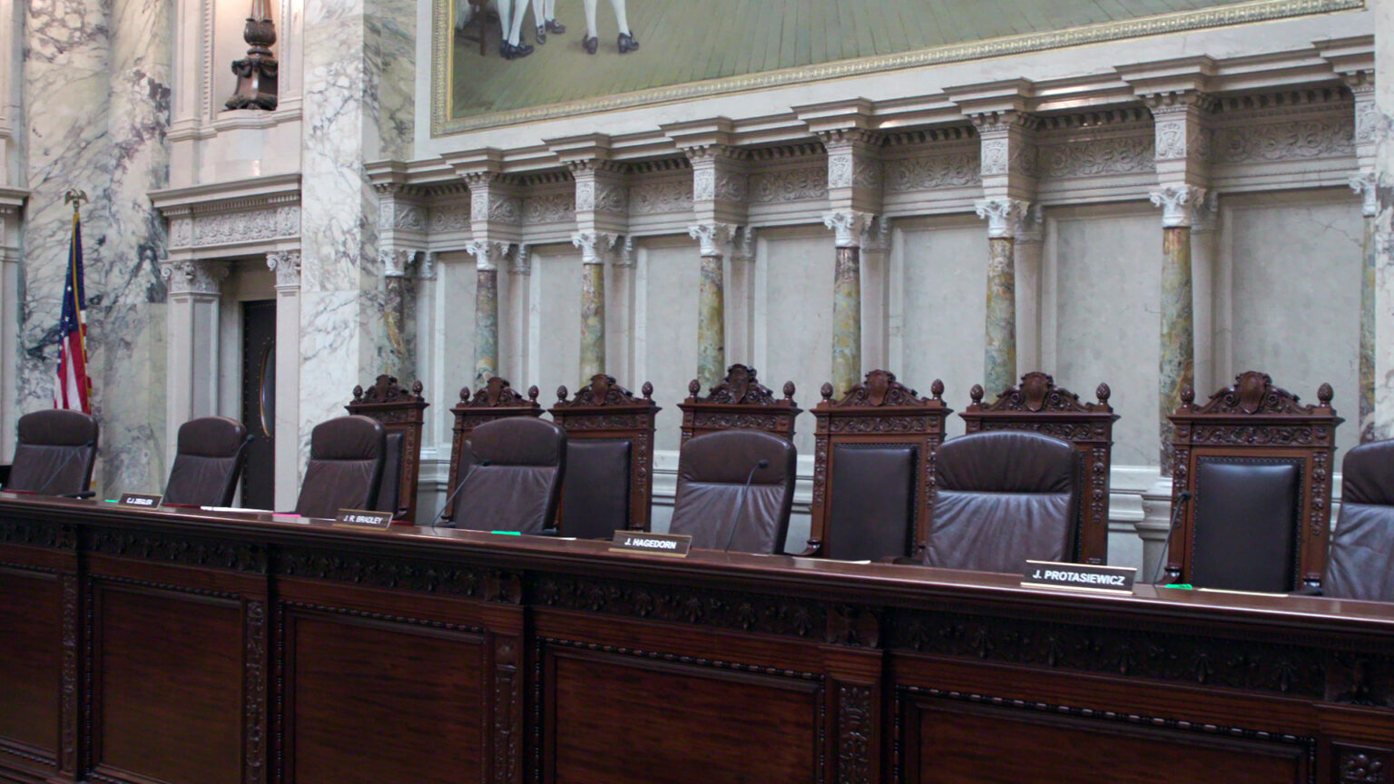 Seven high-backed leather chairs behind a wood bench with nameplates in front of each sit empty, with another row of high-backed wood and leather chairs in the background, in a room with marble masonry, pillars, a large painting and a U.S. flag.