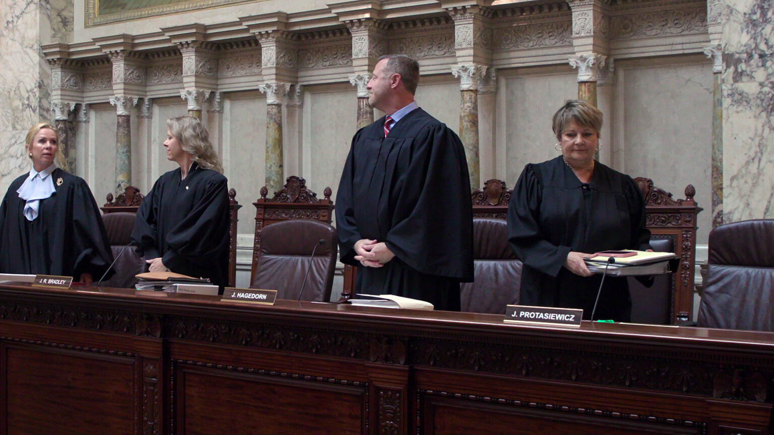 Annette Ziegler, Rebecca Bradley and Brian Hagedorn stand behind a judicial dais and look at each other while Janet Protasiewicz prepares to set down a binder with documents, with one row of high-backed leather chairs behind them and another row of high-backed wood and leather chairs in the background, in a room with marble masonry pillars.