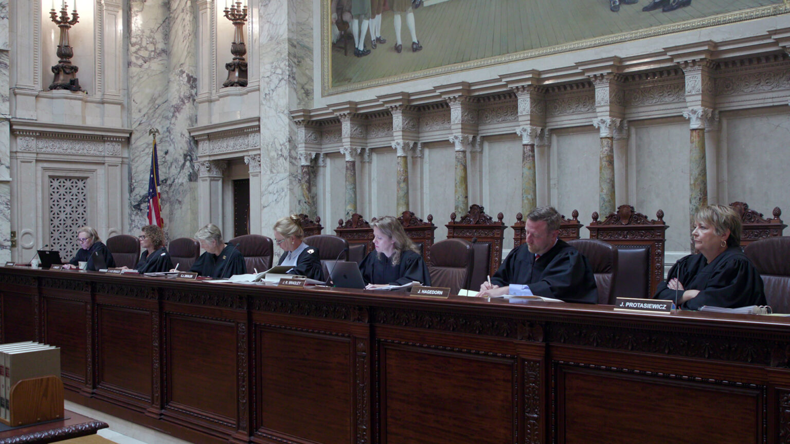 From left to right, Jill Karofsky, Rebecca Dallet, Ann Walsh Bradley Annette Ziegler, Rebecca Bradley, Brian Hagedorn and Janet Protasiewicz sit at a judicial dais in a row of high-backed leather chairs behind them, with another row of high-backed wood and leather chairs behind them, in a room with marble masonry, wall-mounted metal light fixtures, a U.S. flag and a large painting.