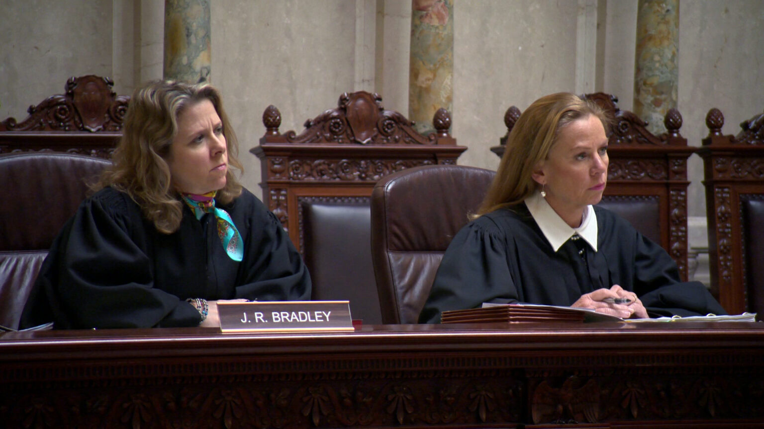 Rebecca Bradley and Annette Ziegler sit in high-backed leather chairs at a judicial bench with a nameplate reading J.R. Bradley and an expanding file folder on its surface, with a row of empty high-backed wood and leather chairs behind them in a room with marble masonry.