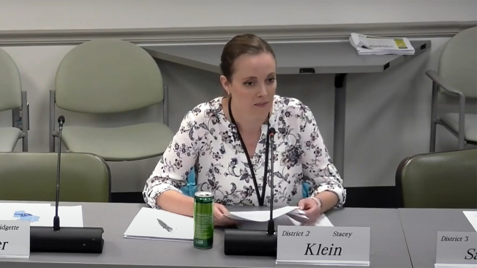 Stacey Klein sits at a table and speaks into a microphone while holding a piece of paper, with a nameplate in front of her reading District 2 and Stacey Klein, empty chairs on either side of her, and more empty chairs placed against a wall in the background.
