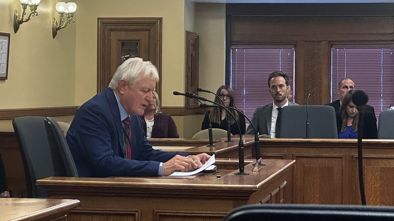 Jim VandenBrook sits in a wood witness stand mounted with multiple microphones and speaks while looking at a sheet of paper, with people seated in the background in a wood gallery space, in a room with brass light fixtures and wood-framed windows with closed blinds.