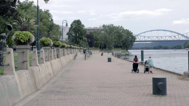 Two people push strollers along a brick-paved walkway between a river and a concrete levee, with trees and other plants growing alongside lawns and buildings on the upper level, and with two bridge spans and bluffs on the opposite side of the water in the background.