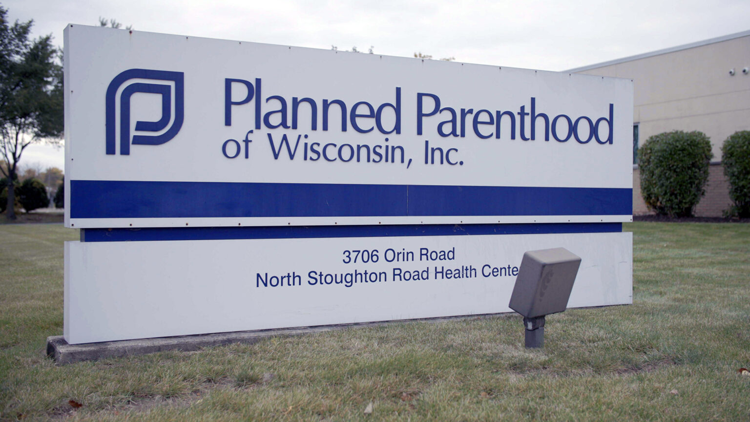 A sign reading Planned Parenthood of Wisconsin, Inc. with an address stands in front of a single-story building surrounded by a lawn, bushes and trees.