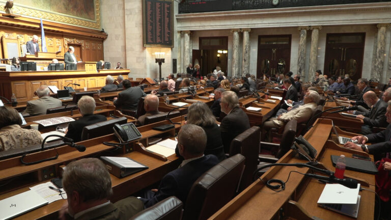Legislators sit in curved rows of wood desks equipped with microphones and phones, and face a legislative dais where multiple people sit and stand, in a room with marble masonry pillars and tall double doors with oval windows.