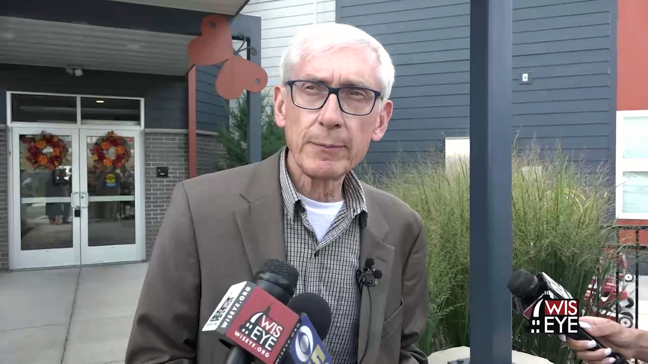 Tony Evers stands outside and speaks into multiple microphones held by other people, with the double-doors of a building entrance in the background.
