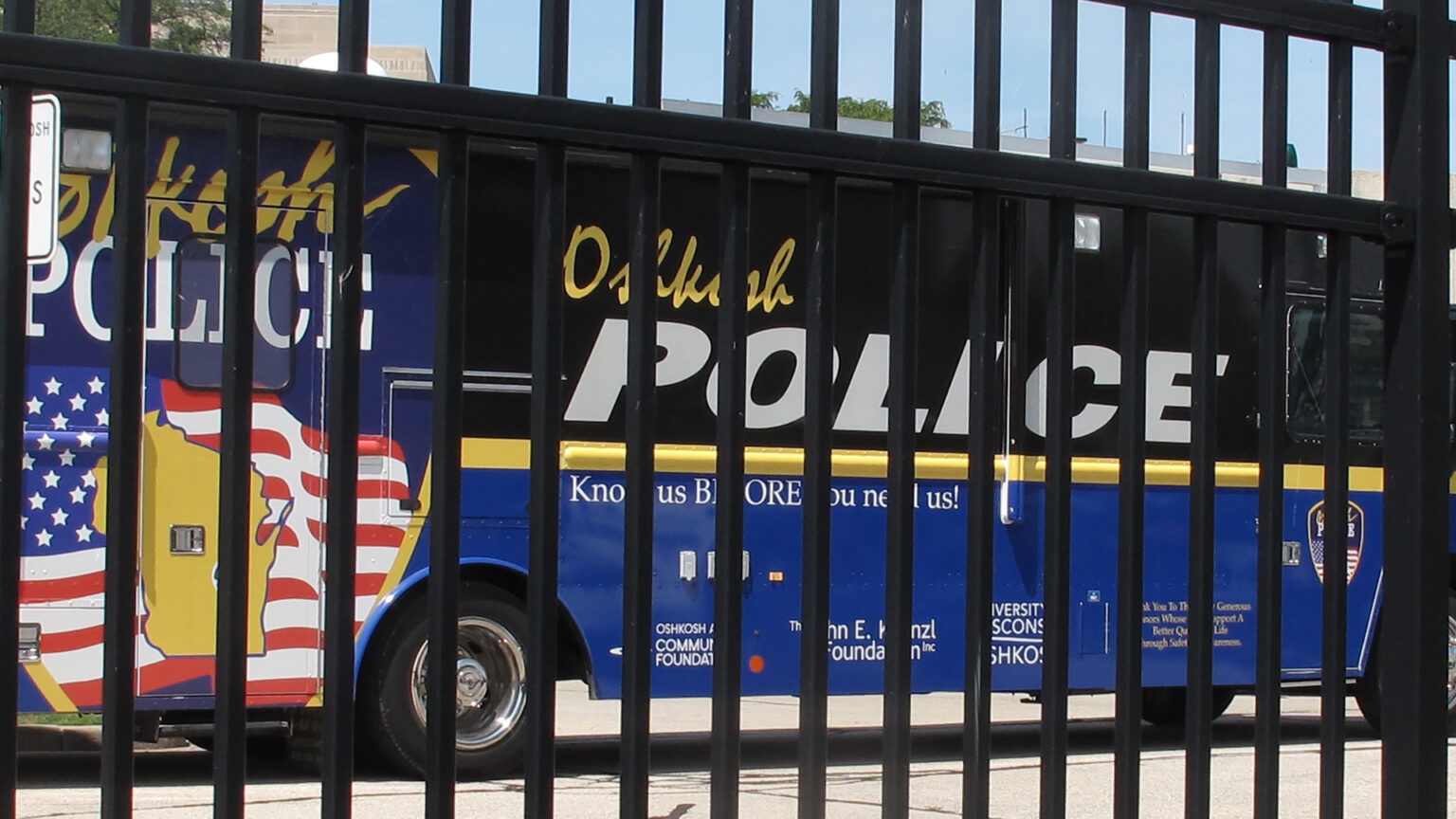 A bus with wrap graphics bearing multiple Oshkosh Police wordmarks, a graphic showing an outline of the state of Wisconsin atop the U.S. flag and other graphics is seen parked behind a painted metal fence with vertical bars.