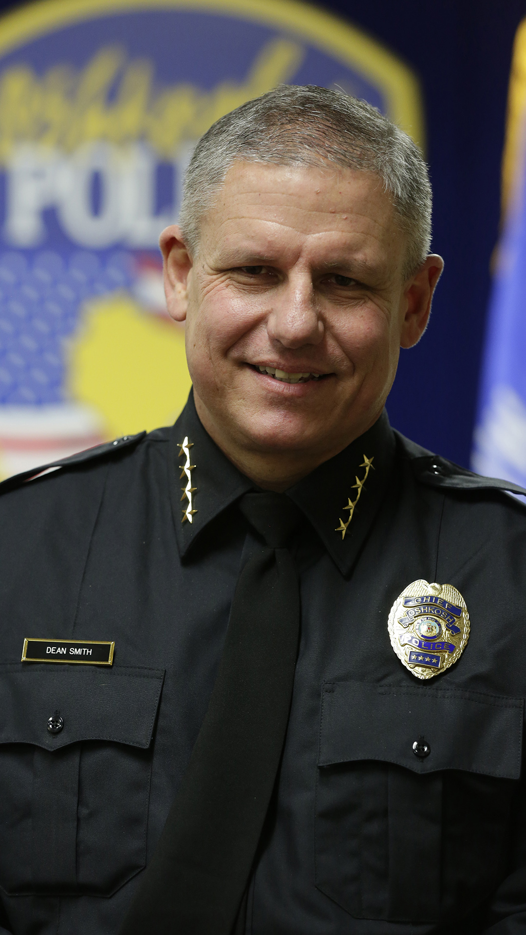 Dean Smith poses for a portrait while wearing a police officer's uniform in front of an out-of-focus Oshkosh Police badge graphic.