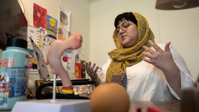 Hanan Jabril gestures with both hands while speaking and seated in an office with multiple items on a desk in the foreground and various hand-drawn illustrations and photos attached to a wall in the background.