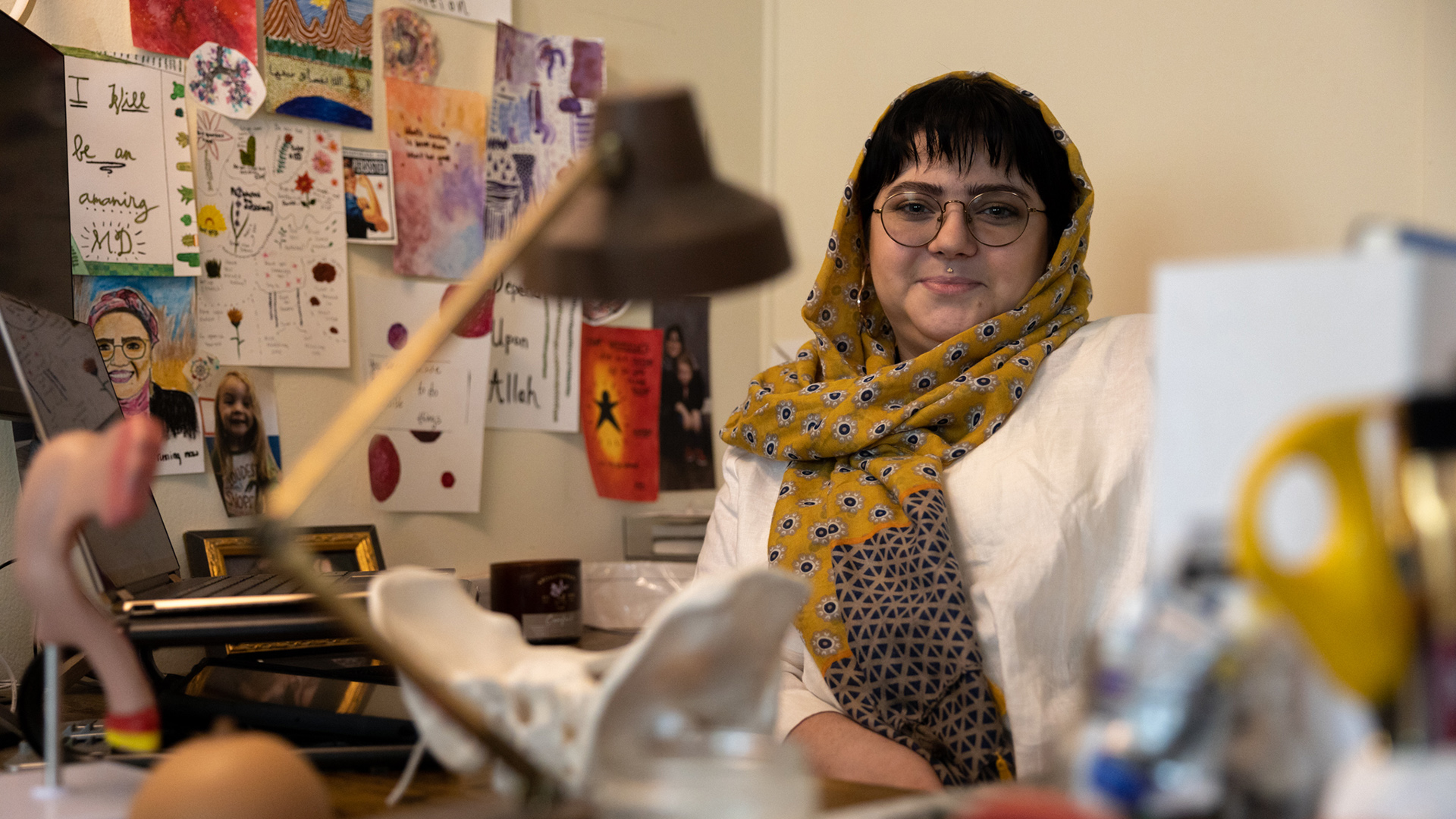 Hanan Jabril poses for a portrait while sitting in an office with multiple items on a desk in the foreground and various hand-drawn illustrations and photos attached to a wall in the background.