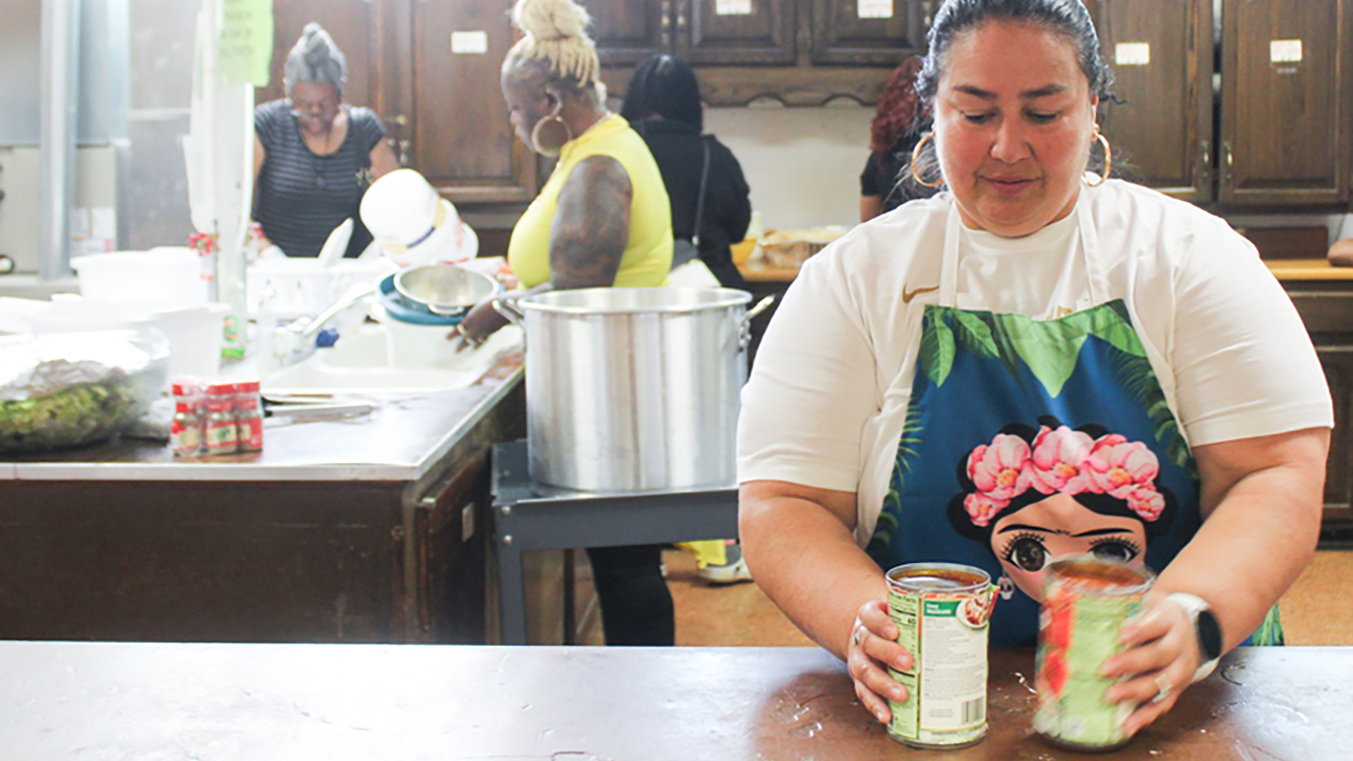 Maria Beltran holds two open items of canned food on a countertop in an institutional kitchen space, with other people in the background handling cooking implements and food storage containers on an island countertop and another counter along a row of cabinets in the background.