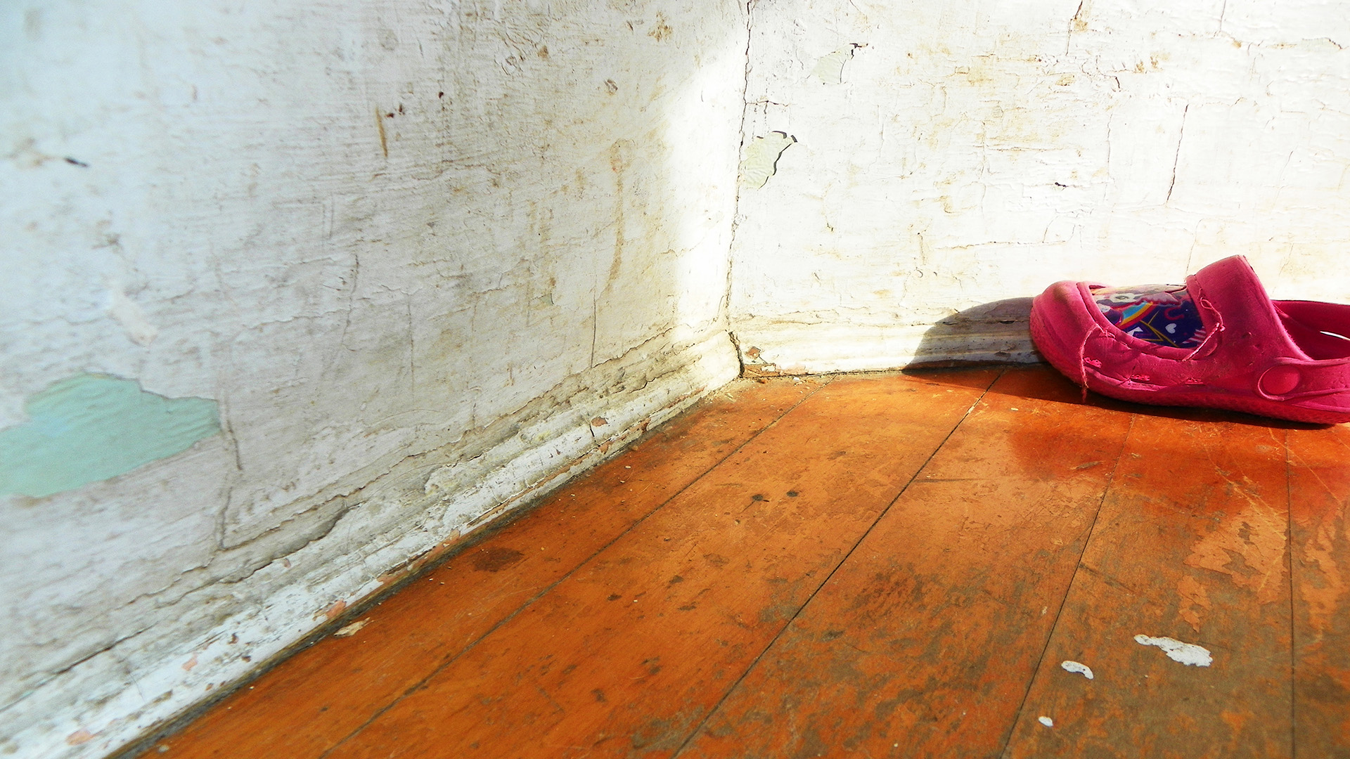 A single shoe sits on a wooden floor next to a corner of two walls with cracked and peeling layers of paint.